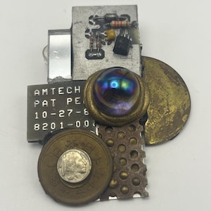 Könnte beinhalten: Eine Brosche aus verschiedenen Materialien, darunter eine Leiterplatte mit elektronischen Bauteilen, eine Metallplatte mit der Aufschrift AMTECH PAT PEN 10-27-8 8201-00, eine zentrale irisierende Glaskugel, eine japanische Münze und eine Messingscheibe.