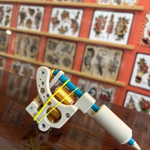 May include: A tattoo machine frame in an off-white matte finish with two gold-coloured coils, secured by a bright yellow rubber band. A blue pencil is inserted through the frame, extending through a matching off-white cylindrical grip at the front.