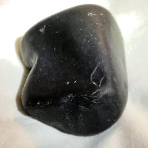 Pode incluir: Uma pedra polida e lisa de cor cinza-carvão escuro com uma forma arredondada e irregular. A superfície apresenta inclusões minerais claras e subtis veios brancos, criando um aspeto natural e manchado sobre a base escura e sólida.