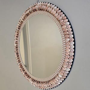 Op de afbeelding: Een ronde wandspiegel met een frame versierd met meerdere lagen kleine, lichtroze spiraalschelpen in een cirkelvormig patroon, omrand door een dik, gedraaid wit touw op een donkere, ronde achtergrond.