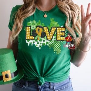 May include: Green t-shirt with a gold "LOVE" graphic featuring shamrocks and a checkered pattern. The graphic also includes a cartoon lineman with the text "Linemen Rock".