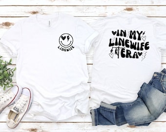 May include: Two white t-shirts with a black graphic design. The front shirt has a smiley face with hearts for eyes and the text "Linewife". The back shirt has the text "In My Linewife Era" with a power line and hearts.