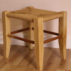 May include: A small stool with four light-colored wooden legs and a rectangular seat woven from natural tan fiber. The legs are connected by thin, dark brown wooden support rungs on all four sides, creating a sturdy frame for the woven seat.