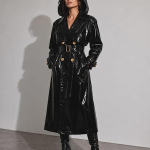 May include: A long, black, glossy patent trench coat featuring a hood, a double-breasted front with gold-toned buttons, and a matching belt with a rectangular buckle. The coat has long sleeves with buckled cuffs and reaches below the knees.