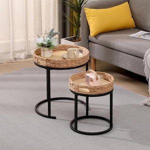 Pode incluir: Um conjunto de duas mesas de apoio com estruturas de metal preto e tampos circulares de madeira rodeados por bordas de erva marinha trançada. A mesa maior contém uma caneca de cerâmica branca e um vaso, a menor contém um rádio rosa.
