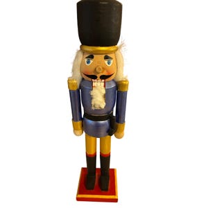 May include: A wooden nutcracker figure wearing a tall black hat, a blue jacket with gold accents, and black boots. The figure has white hair, a painted face with a mustache, and stands on a square red base with a yellow border trim.