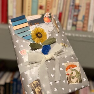 May include: A rectangular package wrapped in grey paper with a white heart pattern, tied with a cream-coloured ribbon. It features a blue wax seal, a yellow sunflower, a set of blue colour swatches, a metallic pen, and stickers of a dog and a frog.