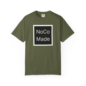 May include: An olive green short-sleeve t-shirt featuring a central graphic design. The design consists of a black square with a white border, containing the white text NoCo Made centered in a clean, sans-serif font on the front of the shirt.