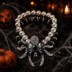 May include: A stretch bracelet featuring alternating metallic gold-tone spherical beads and faceted black beads, centered with a large, detailed spider charm. The spider is embellished with small clear crystals and a central black stone on its back.