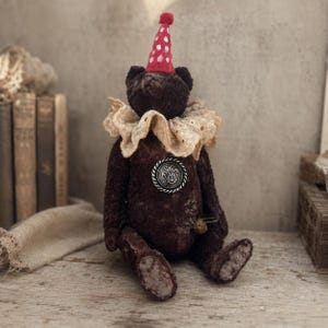 May include: A dark brown teddy bear wearing a red party hat with white polka dots and a beige ruffled lace collar. The bear features a silver circular button on its chest with an embossed crest and a small safety pin attached to its torso.