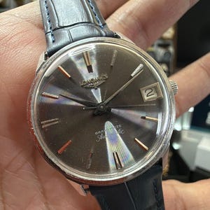 May include: A Longines wristwatch with a circular silver-tone case and a dark grey sunburst dial. The watch features silver-tone hour markers, a date window at the three o'clock position, and a black leather strap with a textured crocodile pattern.