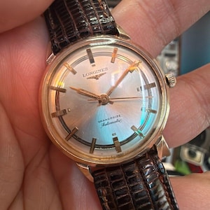 May include: A gold-tone Longines Grand Prize Automatic wristwatch with a silver dial, gold-tone hour markers, and a dark brown textured leather strap. The watch face features the Longines logo, the text Grand Prize, and the word Automatic.