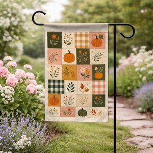 May include: A rectangular garden flag featuring a patchwork grid design with alternating squares of pumpkins, autumn flowers, foliage, and plaid patterns in shades of orange, green, cream, and brown, suspended from a black metal stand.