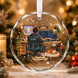 May include: A round, faceted glass Christmas ornament featuring a printed illustration of a person sitting at a desk with a computer, a dog in a bed, and the text 'OLIVER', '2026', and 'Merry Christmas'. A white ribbon is attached to the top.