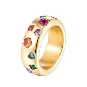 Op de afbeelding: Een brede goudkleurige ring met een glad, gepolijst oppervlak, ingelegd met meerdere kleine, gefacetteerde edelstenen in diverse kleuren zoals roze, paars, oranje, blauw en groen, gerangschikt in een onregelmatig, verspreid patroon.