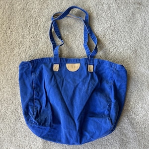 May include: A royal blue canvas tote bag featuring two shoulder straps and a semi-circular tan leather patch centered at the top. The patch is embossed with the text GITANO. The bag has a wide, open main compartment and reinforced stitching.