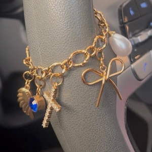 May include: A gold-toned chain charm bracelet featuring a variety of dangling pendants including a gold wing, a round blue gemstone, a crystal-encrusted cross, a simple gold bow, and a single white teardrop-shaped pearl charm.