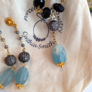 May include: A pair of earrings with a blue gemstone, a silver bead, and a gold bead. The earrings are on a white fabric background. The fabric has the text "Porcelain Smith" printed on it.