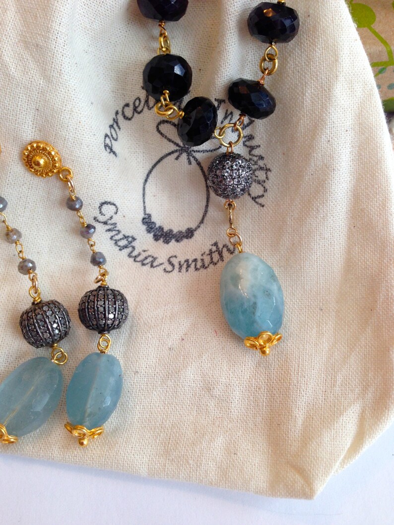 May include: A pair of earrings with a blue gemstone, a silver bead, and a gold bead. The earrings are on a white fabric background. The fabric has the text "Porcelain Smith" printed on it.