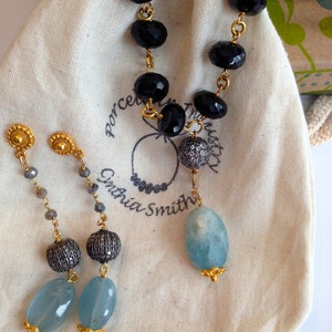 May include: A pair of earrings with a blue gemstone, a silver bead, and a gold chain. The earrings are displayed on a white fabric bag with the text "Porcelain Trust" and "Cynthia Smith" printed on it. The bag also contains a necklace with black beads and a blue gemstone.