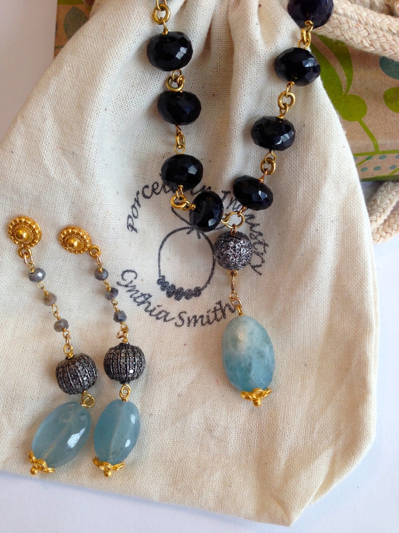 May include: A pair of earrings with a blue gemstone, a silver bead, and a gold chain. The earrings are displayed on a white fabric bag with the text "Porcelain Trust" and "Cynthia Smith" printed on it. The bag also contains a necklace with black beads and a blue gemstone.