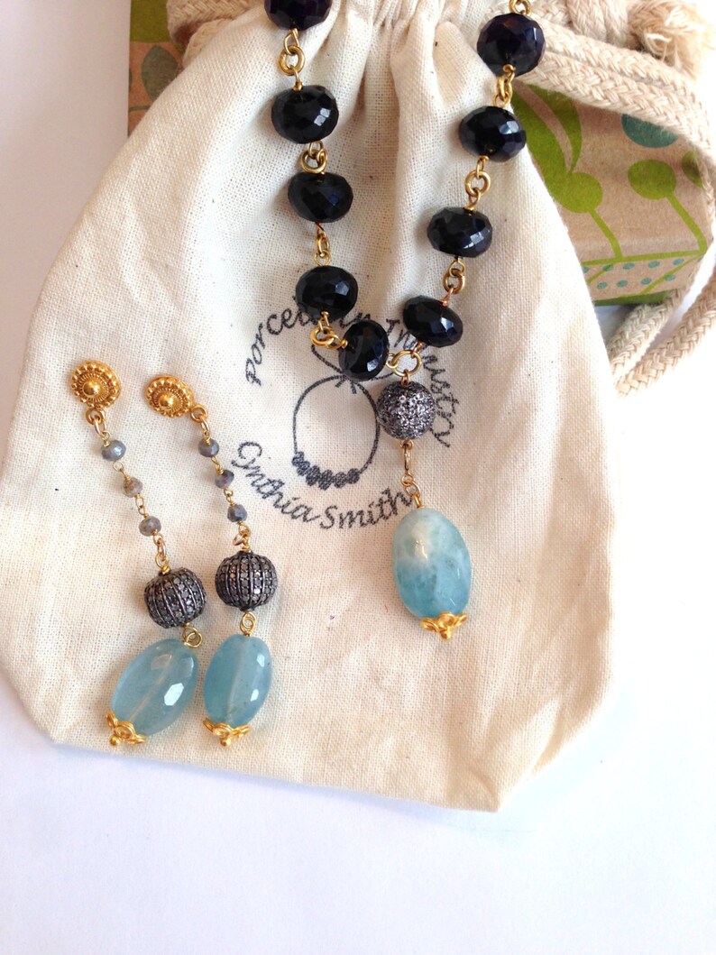 May include: A necklace and earrings set featuring black onyx beads, aquamarine stones, and gold accents. The necklace is in a small white drawstring bag with the text "Porcelain Trust" and "Cynthia Smith" printed on it.