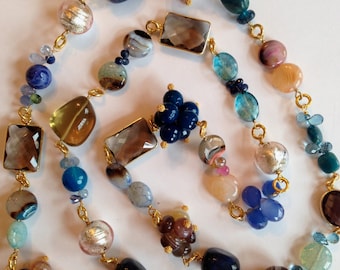 May include: A long necklace with a variety of colorful gemstone beads, including blue, green, brown, and pink. The necklace is made with gold-toned metal.