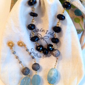 May include: A set of black onyx and aquamarine earrings with gold accents. The earrings are on a white fabric background with a small bag and the text "Cynthia Smith".