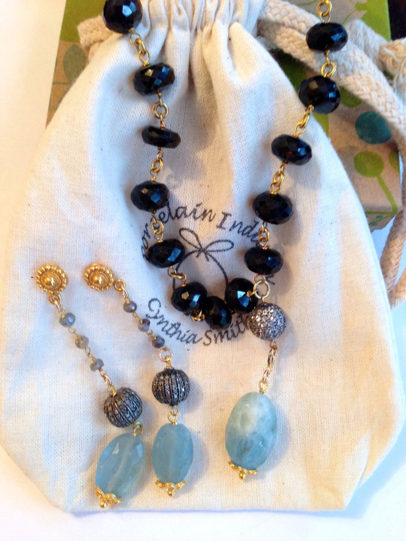 May include: A set of black onyx and aquamarine earrings with gold accents. The earrings are on a white fabric background with a small bag and the text "Cynthia Smith".