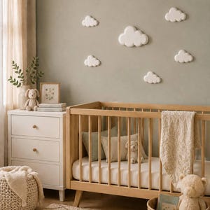 Puede incluir: Una cuna de madera clara con barrotes verticales, un colchón blanco y una manta beige texturizada. La pared presenta cinco decoraciones de pared blancas con forma de nube. También hay una cómoda blanca de tres cajones, una cesta y peluches.