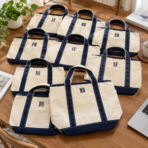 Op de afbeelding: Een collectie van acht off-white canvas draagtassen met marineblauwe randen en hengsels. Elke tas is voorzien van een voorvak en is geborduurd met unieke monogrammen van twee letters, waaronder EE, MB, BW, PM, CC, NF, NS, DN en MO.