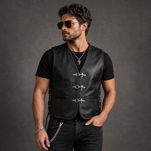 Könnte beinhalten: Eine schwarze Lederweste mit drei silberfarbenen Metallhakenverschlüssen auf der Vorderseite, geflochtenen Lederkanten und seitlichen Schnürdetails. Die Weste wird über einem schlichten schwarzen T-Shirt mit einer silbernen Kettenhalskette getragen.