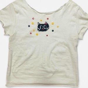 May include: A cream-colored short-sleeve t-shirt featuring an embroidered black cat face with white eyes and a red nose. The design is surrounded by small, colorful embroidered stars, hearts, and circular buttons in red, yellow, and dark blue hues.