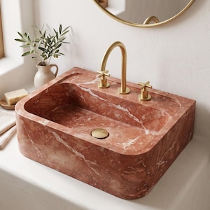 May include: A rectangular bathroom sink crafted from reddish-brown marble with white veining. The sink features a brushed gold faucet with two cross-handle controls and a matching gold drain cover, set against a smooth, light-colored countertop.