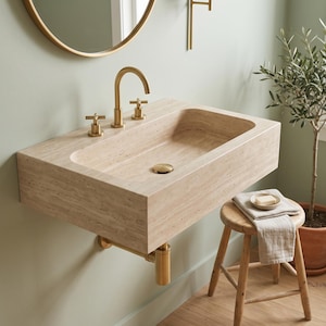 May include: A wall-mounted rectangular bathroom sink crafted from beige travertine stone, featuring a polished brass faucet with two cross-handle knobs and a matching brass drain stopper, mounted against a light sage green wall with visible plumbing.