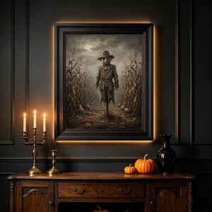 May include: A framed wall art print depicting a scarecrow standing in a cornfield under a moonlit sky. The artwork features muted brown and grey tones, showcasing a textured scarecrow figure amidst tall stalks of corn and scattered pumpkins.