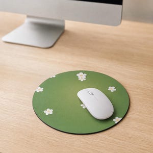 May include: A round mouse mat with a solid green background featuring a repeating pattern of small white flowers with yellow centres. A white wireless computer mouse is positioned on top of the mouse mat, which rests on a light wood desk surface.