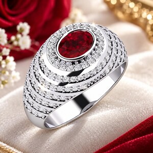 Könnte beinhalten: Ein silberfarbener Ring mit einem zentralen, oval geschliffenen, tiefroten Edelstein. Die Ringschiene ist breit und gewölbt und mit mehreren konzentrischen Reihen kleiner, klarer, funkelnder Steine besetzt, die den roten Stein umgeben.