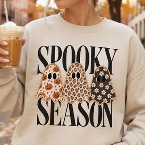 Może przedstawiać: Beżowa bluza z okrągłym dekoltem, czarnym napisem SPOOKY SEASON i trzema ilustracjami duchów. Pierwszy duch ma wzór w dynie i gwiazdki, drugi w cętki lamparta, a trzeci w białe kwiaty na czarnym tle.