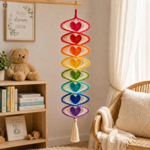 May include: A vertical hanging decoration featuring a series of nine crocheted eye-shaped motifs in rainbow colors, each containing a solid heart in the center. The piece is finished with a cream-colored tassel at the bottom and a gold ring.