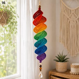 May include: A hanging spiral decoration featuring a vibrant rainbow-colored crochet pattern. The structure transitions from red to orange, yellow, green, blue, and purple. It is accented with wooden beads at the top and bottom, ending in a clear crystal.