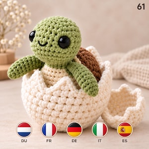 May include: A crocheted turtle toy with a light green head, dark brown shell, and cream-colored body, emerging from a matching cream-colored crocheted eggshell. The turtle has large black plastic eyes and a stitched smile. The number 61 is visible.