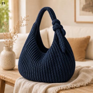 Op de afbeelding: Een donker marineblauwe hobo-handtas gemaakt van dikke, geribbelde gebreide stof. De tas heeft een enkele geïntegreerde schouderband met een opvallend decoratief knoopdetail aan één kant, wat zorgt voor een zacht, afgerond en slungelig silhouet.