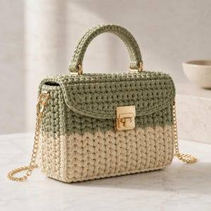 May include: A structured handbag crafted from crocheted yarn featuring a two-tone design with a sage green upper section and a cream-colored lower section. It includes a gold-toned metal clasp closure, a matching top handle, and a gold chain shoulder strap.