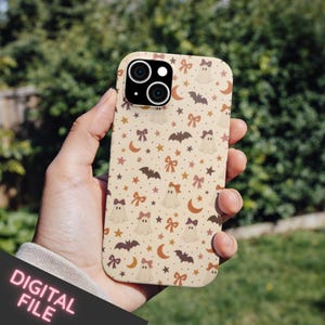 Pode incluir: Uma capa de smartphone com um padrão repetitivo de fantasmas cor de creme com laços, morcegos castanhos, luas crescentes cor de laranja e pequenas estrelas num fundo bege claro. A capa tem um recorte para um sistema de câmara dupla.