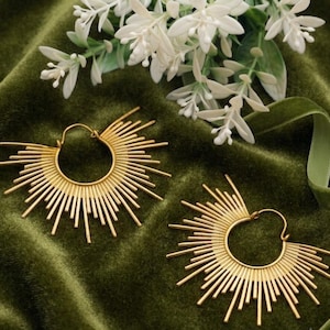 Peut inclure: Une paire de boucles d'oreilles créoles dorées arborant un motif en rayons de soleil avec de fines tiges métalliques rayonnantes partant de la base circulaire. Les boucles d'oreilles présentent une finition polie et un fermoir à tige simple.