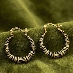 Può includere: Un paio di orecchini a cerchio color oro con un design testurizzato ispirato al bambù. Ogni orecchino circolare presenta un meccanismo di chiusura a cerniera sulla parte superiore e una superficie segmentata e rigata che imita il bambù.
