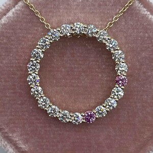 May include: A gold chain necklace featuring a circular pendant set with a continuous row of round brilliant-cut clear stones and two round brilliant-cut pink stones positioned at the bottom right of the circle, resting on a velvet surface.