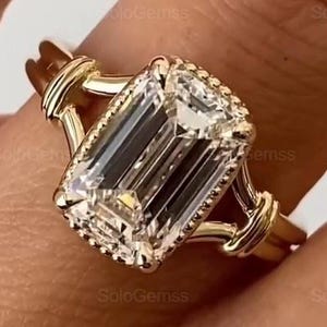 Könnte beinhalten: Ein Goldring mit einem großen, rechteckigen, im Smaragdschliff facettierten Mittelstein. Die Fassung umfasst einen perlenartigen Rand um den Stein und ein geteiltes Ringschienen-Design, bei dem sich das Band in zwei dünne, polierte Golddrähte teilt.