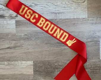 May include: A red satin sash with gold lettering that reads "USC BOUND" and a gold hand symbol. The bottom right corner of the sash has the text "Sassy Sash" in a script font.